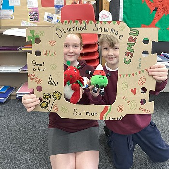 A boy and girl holding a poster that says Diwrnod Shwmae while holding Mr Urdd and the Welsh Dragon