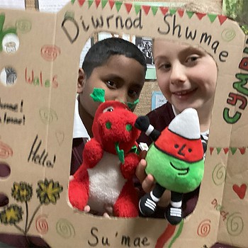Two children holding a poster that says Diwrnod Shwmae while holding Mr Urdd and the Welsh Dragon