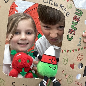 Children holding a poster that says Diwrnod Shwmae while holding Mr Urdd and the Welsh Dragon