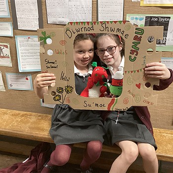 Two girls holding a poster that says Diwrnod Shwmae while holding the Welsh Dragon and Mr Urdd